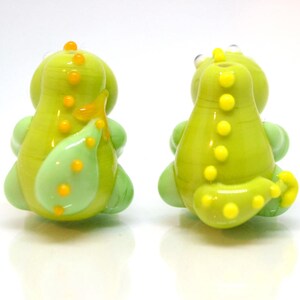 May include: Two green glass dinosaur beads with yellow accents. The beads are about 1 inch tall and have a smooth, glossy finish.