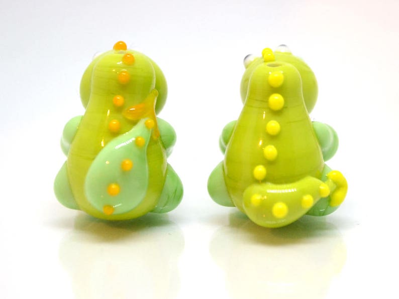 May include: Two green glass dinosaur beads with yellow accents. The beads are about 1 inch tall and have a smooth, glossy finish.