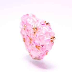 May include: A pink glass bead with a floral design and gold accents. The bead is shaped like a heart and has a delicate, feminine look.