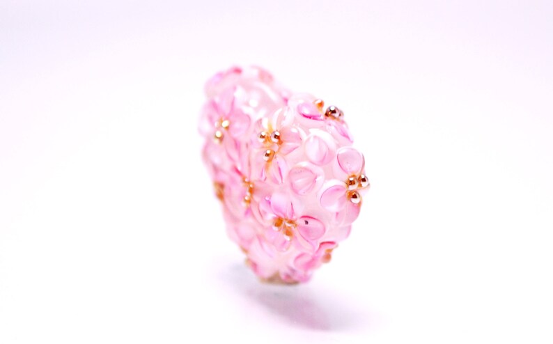 May include: A pink glass bead with a floral design and gold accents. The bead is shaped like a heart and has a delicate, feminine look.