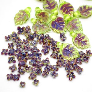May include: A collection of iridescent glass beads in the shape of leaves and flowers. The leaves are green with a purple iridescent sheen, and the flowers are purple with a green iridescent sheen.