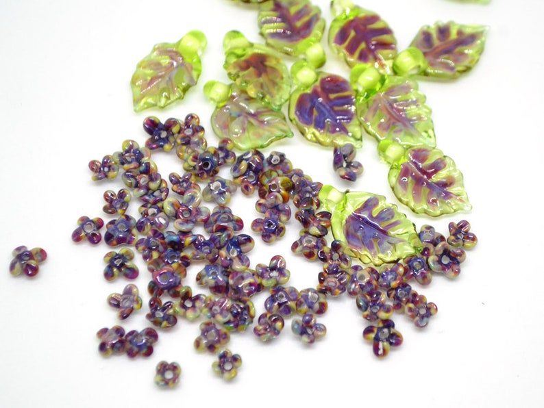 May include: A collection of iridescent glass beads in the shape of leaves and flowers. The leaves are green with a purple iridescent sheen, and the flowers are purple with a green iridescent sheen.
