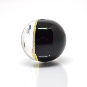 May include: A black and white glass bead with a gold stripe. The bead is round and has a clear glass section with white and blue flecks.