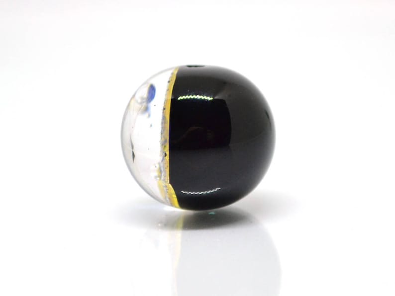 May include: A black and white glass bead with a gold stripe. The bead is round and has a clear glass section with white and blue flecks.