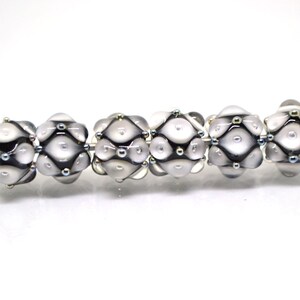 May include: Six white glass beads with black and silver accents. The beads have a textured surface and are arranged in a row.