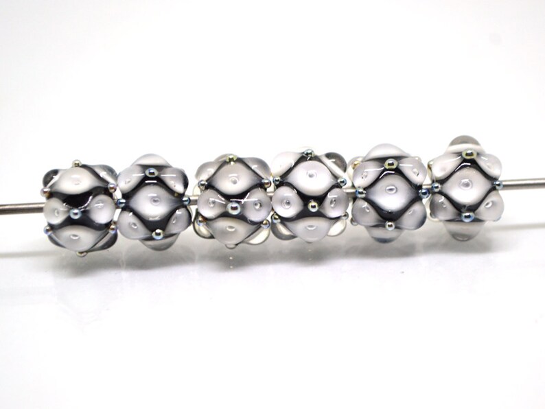 May include: Six white glass beads with black and silver accents. The beads have a textured surface and are arranged in a row.