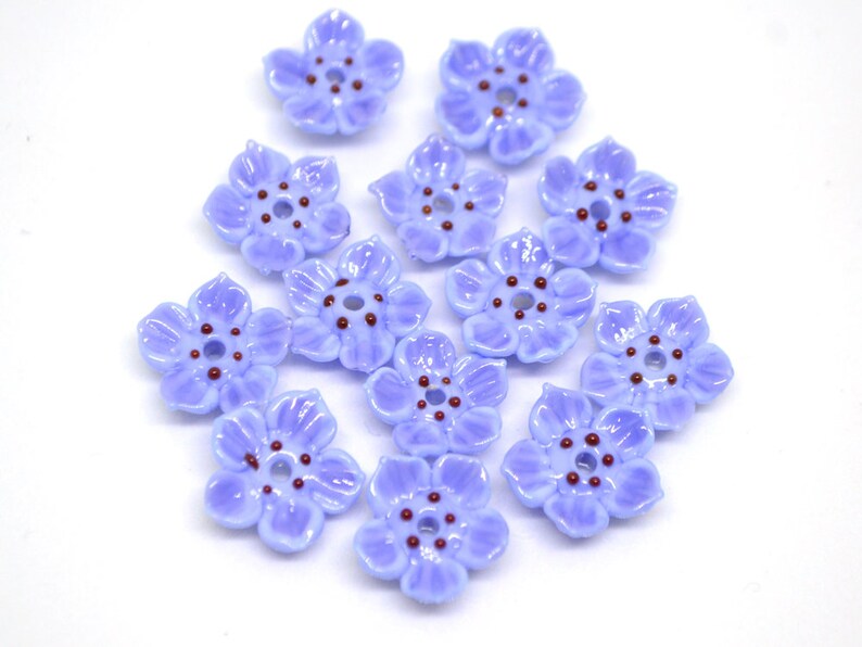 May include: A collection of 12 light blue flower-shaped glass beads with a brown center. The beads are arranged in a circle on a white background.