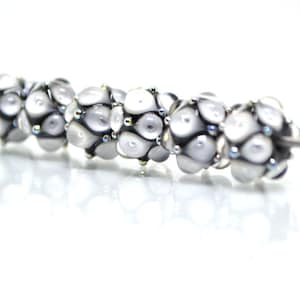 May include: A string of six white glass beads with black and silver accents. The beads are round and have a textured surface. The beads are strung on a silver wire.