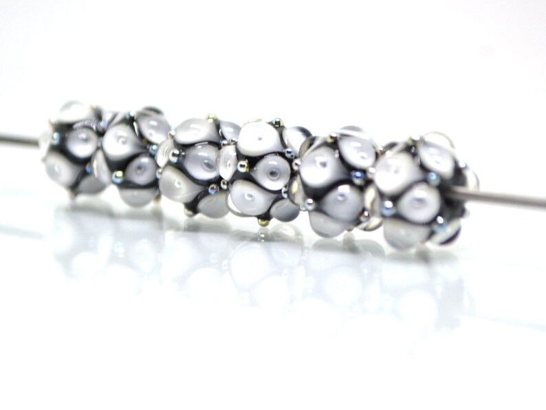May include: A string of six white glass beads with black and silver accents. The beads are round and have a textured surface. The beads are strung on a silver wire.