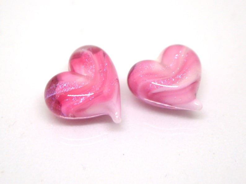 Peut inclure: Deux c&oelig;urs en verre rose avec des tourbillons blancs et des paillettes. Les c&oelig;urs sont de couleur rose clair avec des tourbillons blancs et des paillettes partout.