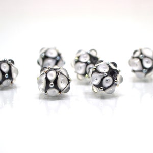 May include: Six white and black glass beads with a geometric pattern. The beads have a shiny, iridescent finish.
