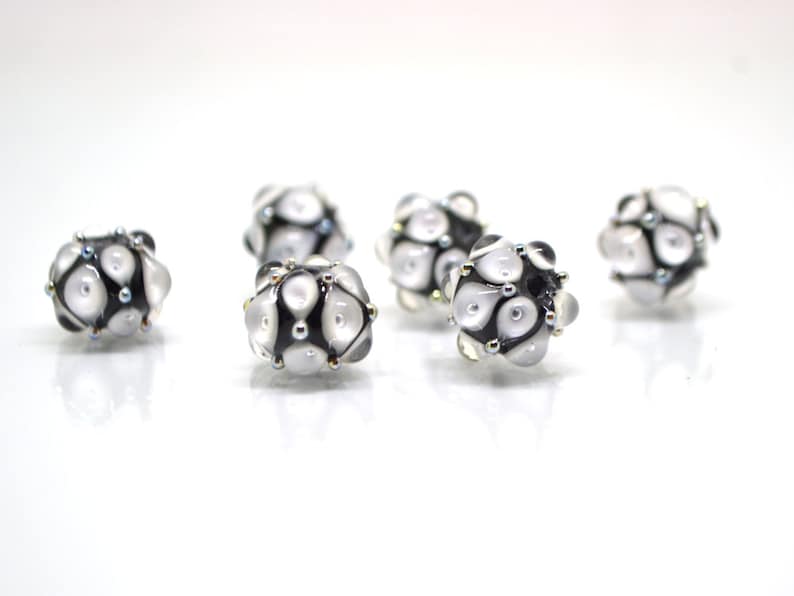 May include: Six white and black glass beads with a geometric pattern. The beads have a shiny, iridescent finish.