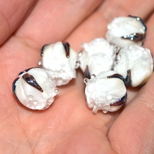 May include: Six white glass beads with iridescent blue and black accents. The beads are round and have a textured surface.