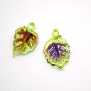 May include: Two green glass leaf charms with purple and brown accents. The leaves have a detailed vein pattern and a hole at the top for hanging.