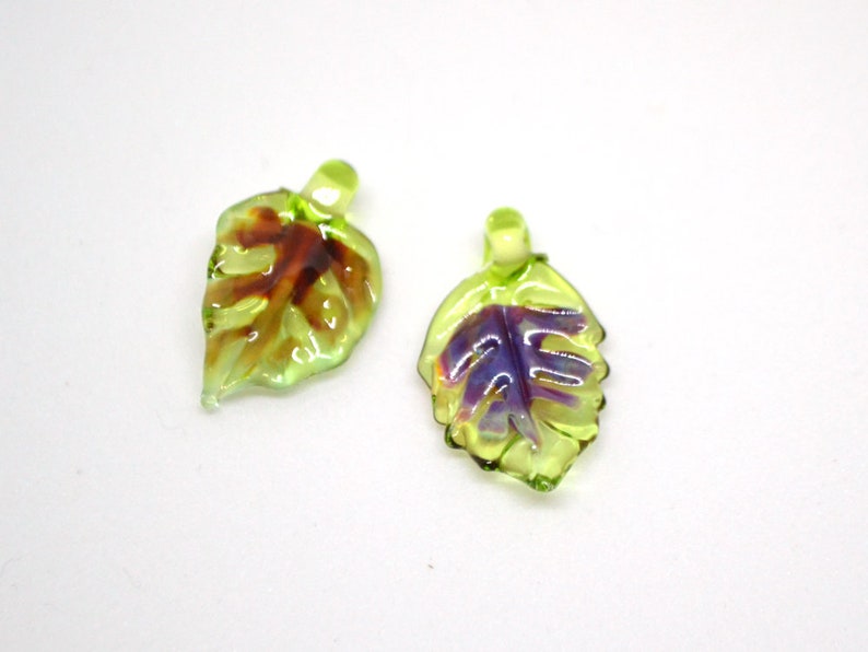 May include: Two green glass leaf charms with purple and brown accents. The leaves have a detailed vein pattern and a hole at the top for hanging.