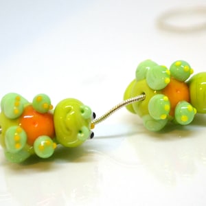 May include: Two green glass beads with orange centers and yellow spots, strung on a silver chain. The beads are shaped like turtles.