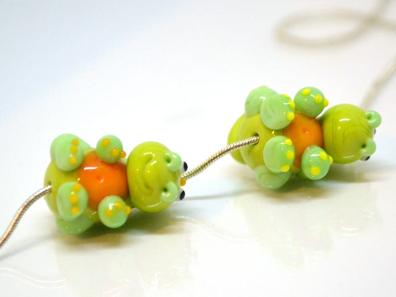 May include: Two green glass beads with orange centers and yellow spots, strung on a silver chain. The beads are shaped like turtles.