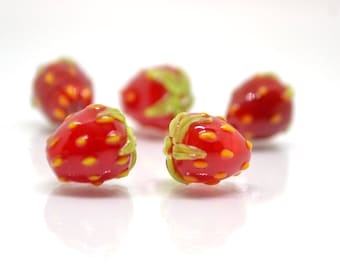 May include: Five glass strawberry beads. The beads are a vibrant red with yellow and orange speckles, and green stems. The strawberries are in focus against a white background, showcasing their glossy texture and detailed craftsmanship.