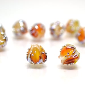 May include: A group of eight glass beads with a unique, textured design. The beads are a mix of orange, yellow, and white with silver accents.