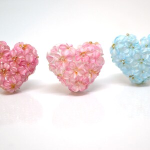 May include: Three heart-shaped floral decorations, one pink, one blue, and one white. Each heart is made of small, delicate flowers.