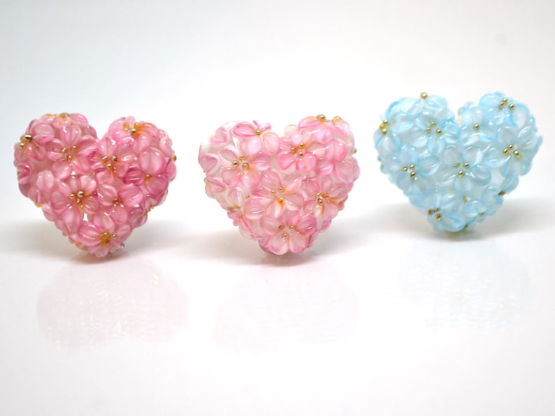 May include: Three heart-shaped floral decorations, one pink, one blue, and one white. Each heart is made of small, delicate flowers.