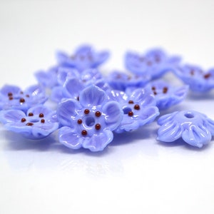 May include: A group of ten light blue glass flower beads with five petals and a brown center. The beads are scattered on a white surface.