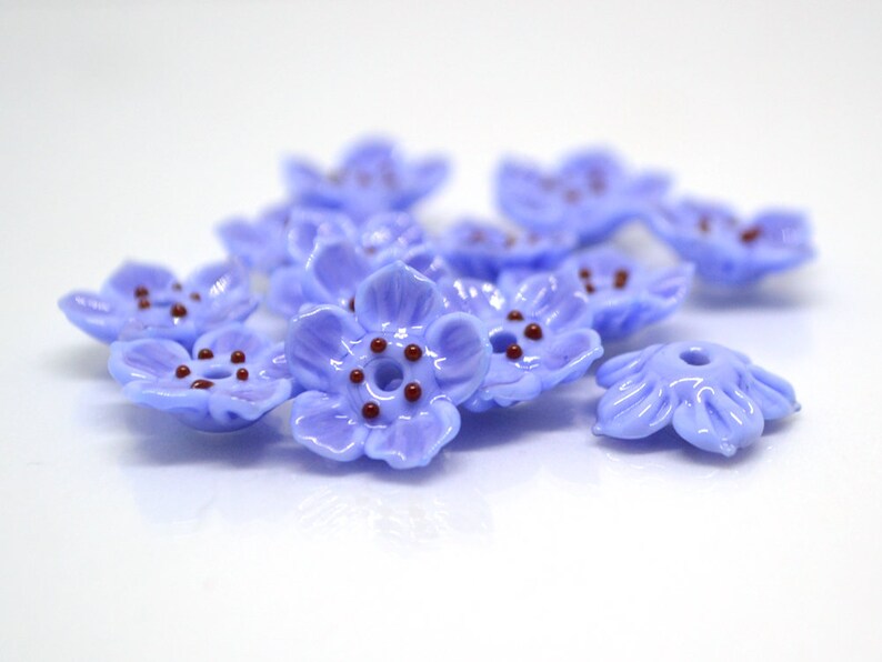 May include: A group of ten light blue glass flower beads with five petals and a brown center. The beads are scattered on a white surface.