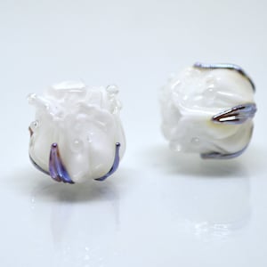 May include: Two white glass beads with a rose-like design and iridescent purple accents.