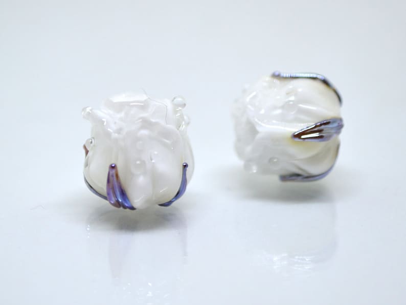 May include: Two white glass beads with a rose-like design and iridescent purple accents.