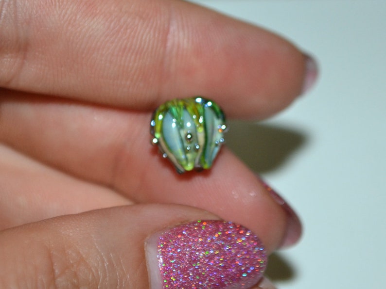 K&ouml;nnte beinhalten: Eine kleine, gr&uuml;ne und wei&szlig;e Glasperle mit einer silbernen Metallkappe. Die Perle hat ein einzigartiges, blumenartiges Design.