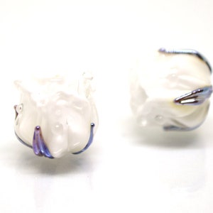 May include: Two white glass beads with iridescent purple and blue accents. The beads are shaped like a flower bud.