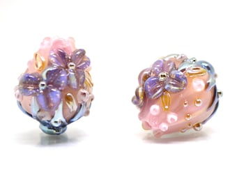 Könnte beinhalten: Zwei rosa Glasperlen mit lila und goldenen Blumen und weißen Punkten. Die Perlen sind irisierend und haben eine strukturierte Oberfläche.