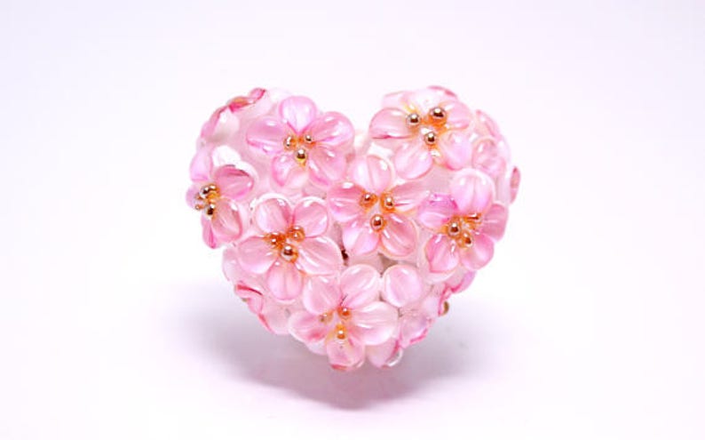 May include: A pink glass heart-shaped ornament with a cluster of pink flowers with gold centers.