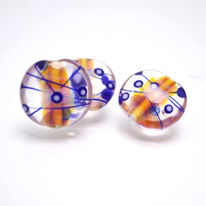 Pu&ograve; includere: Tre perline rotonde di vetro con una base trasparente e disegni astratti in arancio, blu e bianco.