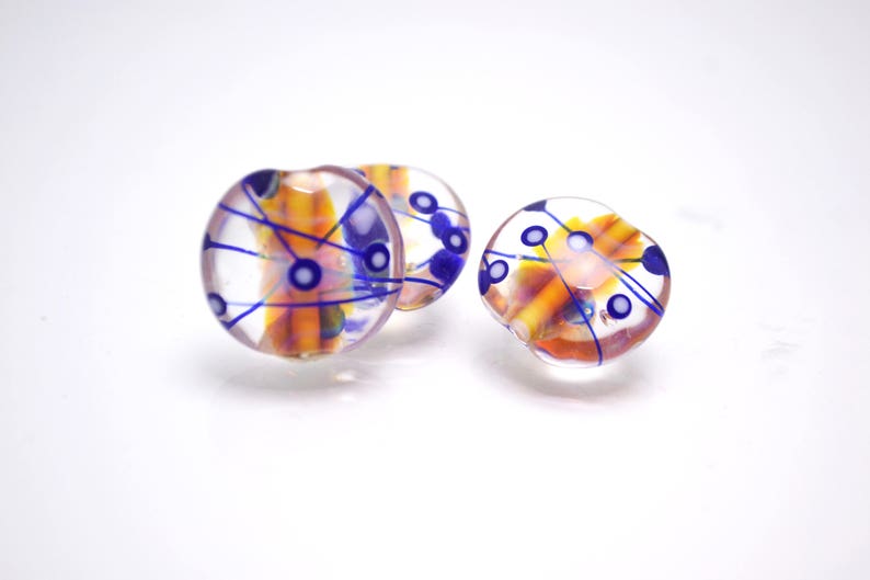 Pu&ograve; includere: Tre perline rotonde di vetro con una base trasparente e disegni astratti in arancio, blu e bianco.