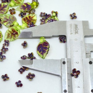 May include: A close-up of a pair of silver calipers measuring a small purple and green glass flower bead. The bead is approximately 2 cm in diameter. The bead is surrounded by other glass beads in the shape of leaves and flowers.