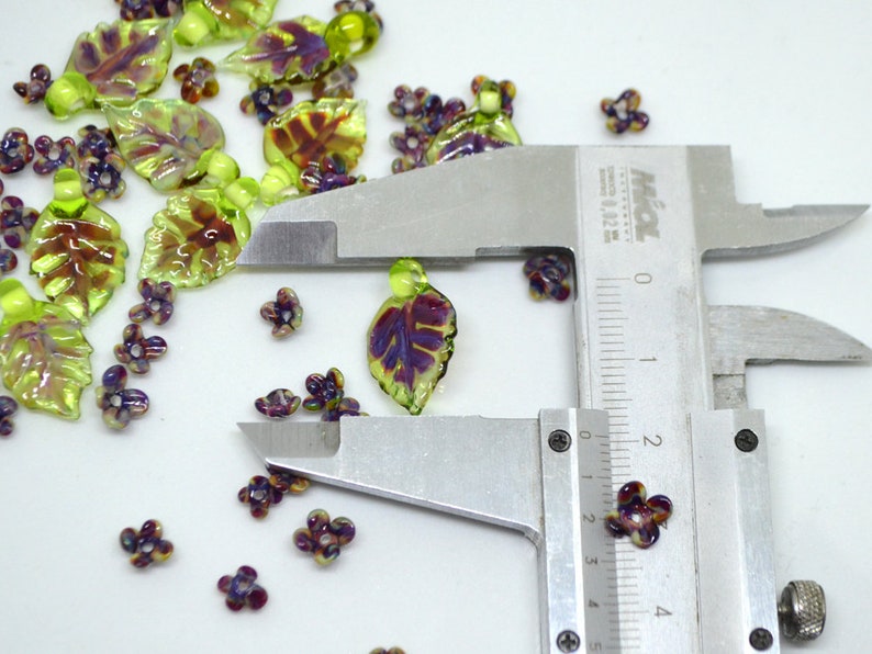 May include: A close-up of a pair of silver calipers measuring a small purple and green glass flower bead. The bead is approximately 2 cm in diameter. The bead is surrounded by other glass beads in the shape of leaves and flowers.