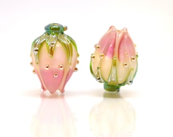 May include: Two glass beads in the shape of a rose bud. The beads are pink and white with green and gold accents.