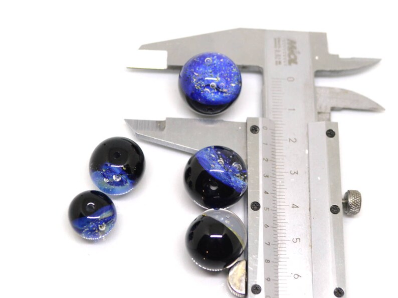 May include: Five blue and black glass beads with silver flecks. The beads are measured with a ruler in inches.