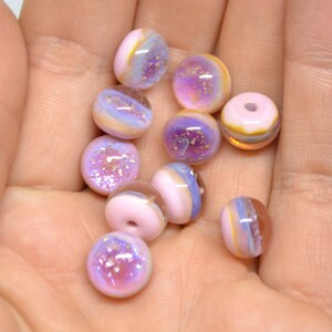 May include: A collection of 12 iridescent glass beads in shades of pink, purple, and white with gold flecks. The beads are round and have a smooth, glossy finish.