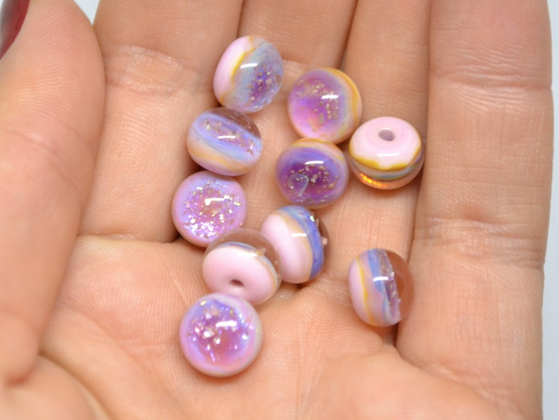 May include: A collection of 12 iridescent glass beads in shades of pink, purple, and white with gold flecks. The beads are round and have a smooth, glossy finish.