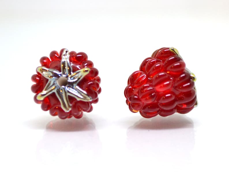 Puede incluir: Dos cuentas de vidrio de color rojo en forma de frambuesa, una con un dise&ntilde;o de estrella dorada y la otra con forma de frambuesa s&oacute;lida.