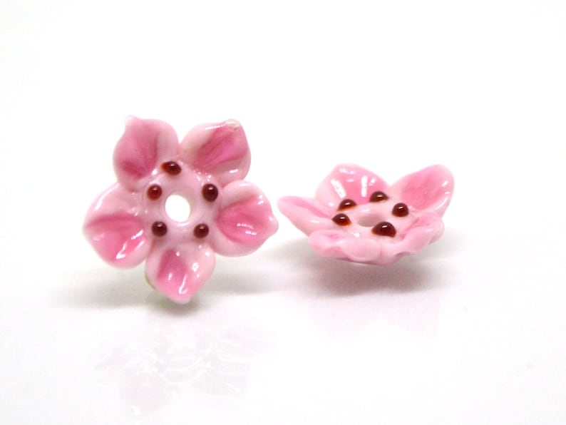 May include: Two pink ceramic flower beads with brown centers. The beads are shaped like five-petal flowers.