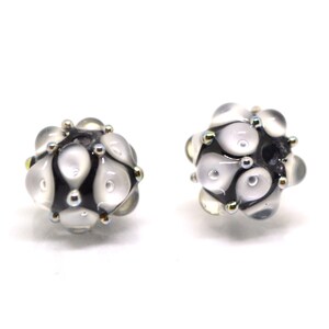 May include: Two white glass beads with black and gold accents. The beads are round and have a textured surface.