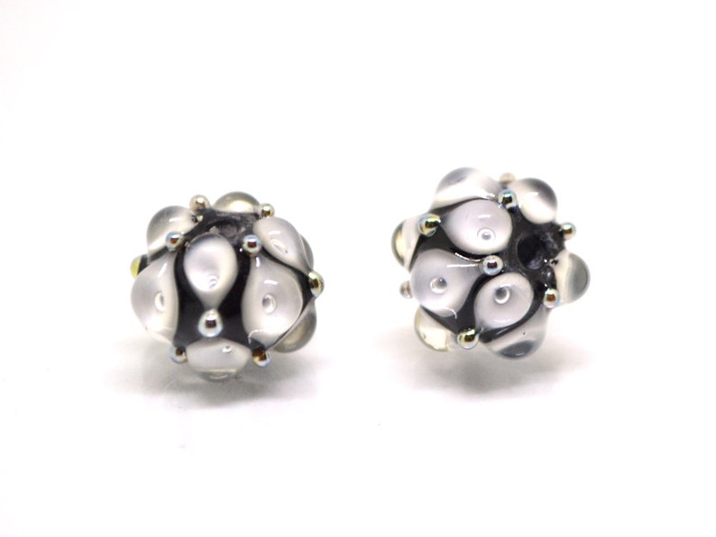 May include: Two white glass beads with black and gold accents. The beads are round and have a textured surface.