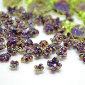 May include: A collection of small, colorful flower-shaped beads. The beads are made of glass and have a variety of colors, including purple, green, and yellow. The beads are scattered on a white surface.