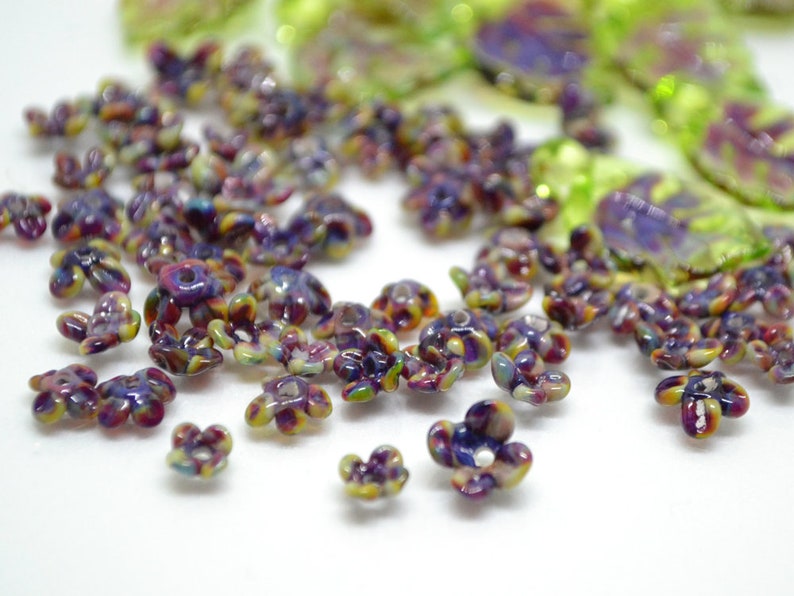 May include: A collection of small, colorful flower-shaped beads. The beads are made of glass and have a variety of colors, including purple, green, and yellow. The beads are scattered on a white surface.