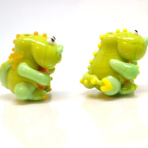 May include: Two small green glass figurines of dinosaurs with yellow accents. The dinosaurs are in a playful pose.
