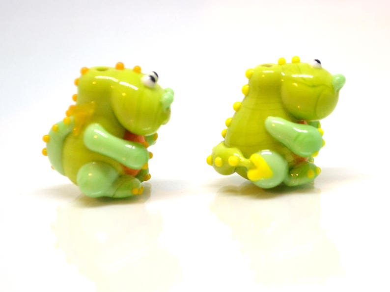 May include: Two small green glass figurines of dinosaurs with yellow accents. The dinosaurs are in a playful pose.
