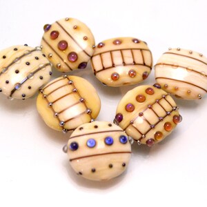 May include: A collection of eight glass beads with intricate patterns and colorful accents. The beads are cream-colored with brown, orange, and blue accents.